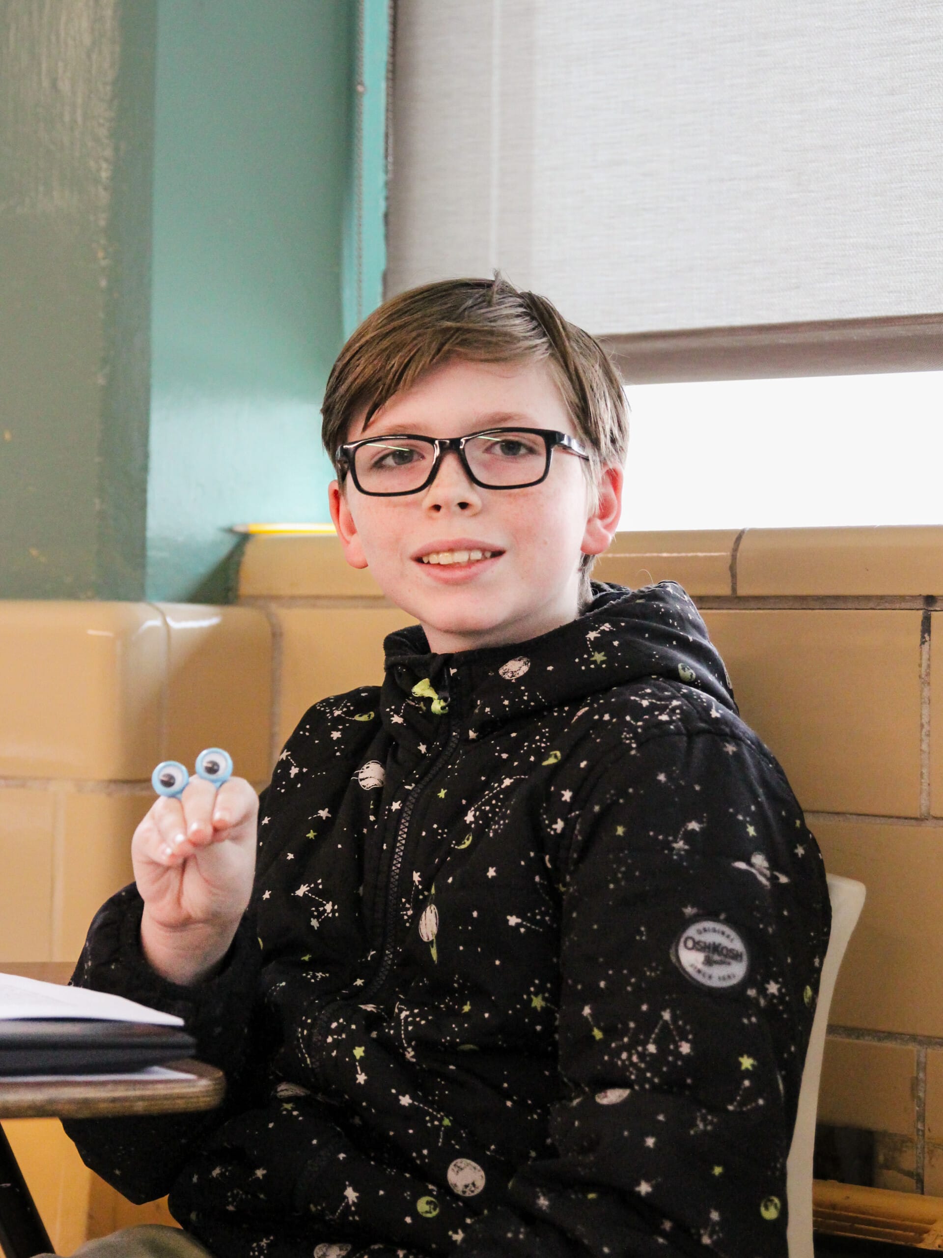 Smiling boy with glasses holding googly eyes in classroom