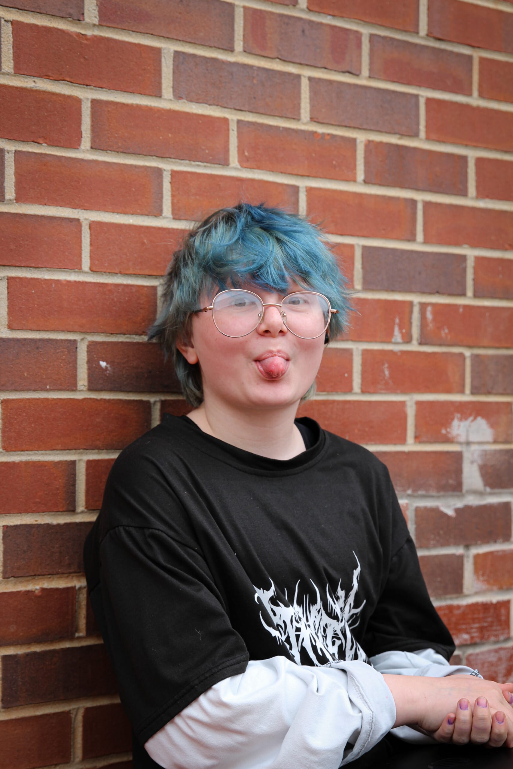 Young person with blue hair sticking tongue out by brick wall