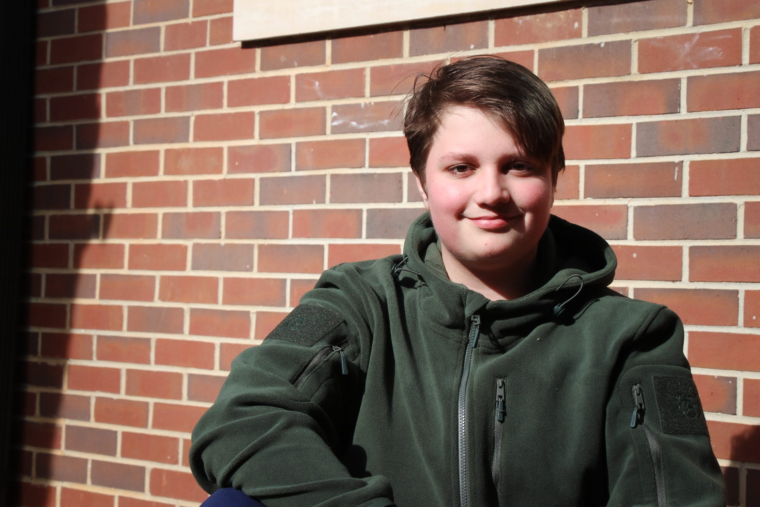 Young person in green jacket smiling against brick wall