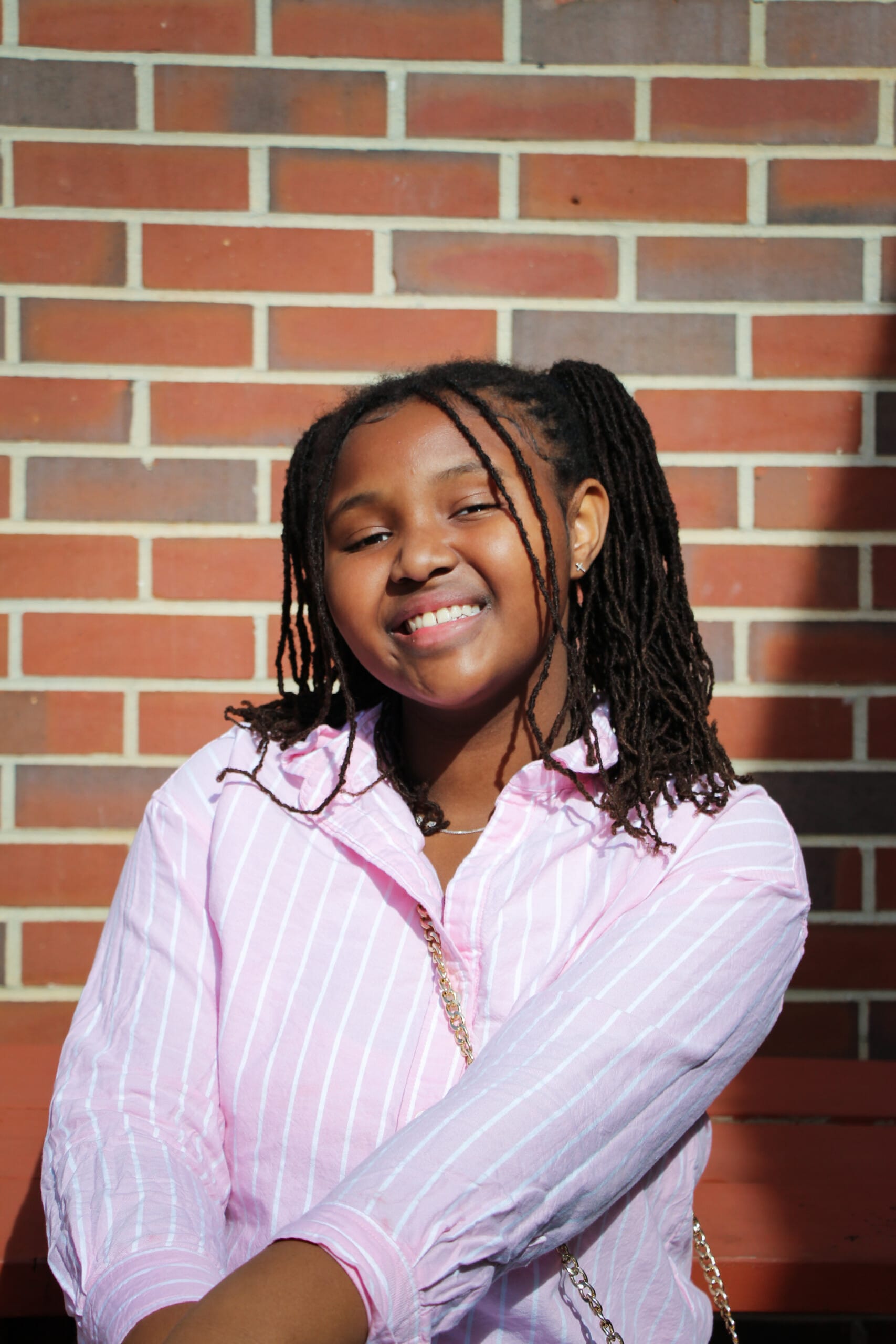 Young girl with locs smiling in pink shirt by brick wall